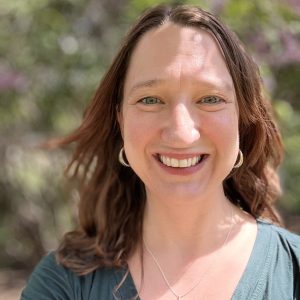 A white woman with brown hair smiling at the camera. She is outside with trees behind her.